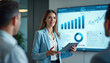 © miss irine - Businesswoman leads corporate seminar for colleagues. Woman presents financial data to coworkers during business meeting. People discuss company strategy using graphs on screen. Speaker with tablet