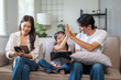 © amnaj - Asian family enjoying time together on couch, father and daughter high fiving, mother using mobile phone