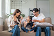 © amnaj - Happy Asian family sitting on couch, using mobile phone and tablet, laughing and spending joyful time