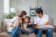 © amnaj - Asian parents and child enjoying time together on living room sofa, mother kissing daughter, father using tablet