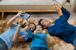 © amnaj - Parents and child lying on living room carpet, using smartphones together, relaxed family bonding over digital content and entertainment