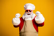 © deagreez - Santa Claus in red outfit poses in a cheerful and festive mood wearing sunglasses and chef hat on a yellow backdrop