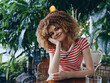© SHOTPRIME STUDIO - Portrait of a joyful young woman with curly hair in a red and white striped tee, sitting at a wooden table amid lush plants and warm natural light.