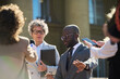 © pressmaster - Middle aged Caucasian woman and young Black man standing outdoors surrounded by journalists holding microphones and smartphones, man gesturing with hand while woman holding folder