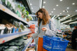 © wifesun - Woman holding a blue shopping basket while meticulously selecting a packaged food item from a refrigerated shelf in a bright supermarket, making conscious choices for healthy living