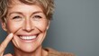 © Tina - Smiling senior Caucasian woman with short hair, wearing a brown sweater. She is pointing at her cheek, showcasing a joyful expression against a gray background.