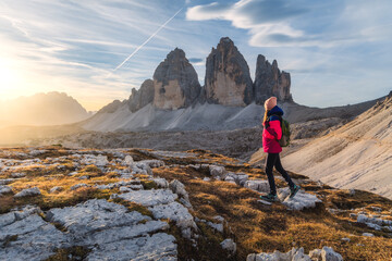 Wall Mural - Girl with backpack on the mountain peak and high rocks at colorful sunset in autumn. Tre Cime, Dolomites, Italy. Landscape with young woman on trail, cliffs, sky with clouds and sun in fall. Hiking