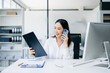 © Nuttapong punna - Asian Business woman Talking on the phone and using a laptop with a smile while sitting at office
