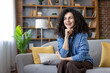 © Liubomir - Young woman with curly hair smiling and daydreaming while relaxing comfortably on a cozy sofa in a modern living room, peaceful and content in casual home surroundings