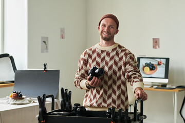 Portrait of man smiling and holding digital camera while standing in photography studio, photo shoot setup and computer monitor with fruit image visible in background