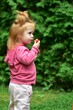 © Karolina - Smiling toddler girl with red hair in a pink hoodie eating a fresh tomato in the garden, enjoying a healthy snack and outdoor play on a bright day surrounded by greenery.