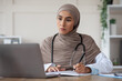 © Prostock-studio - Muslim woman doctor having professional online training on laptop, taking notes while watching webinar, sitting at workdesk in clinic, copy space. Training for doctors, professional development