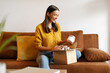 © Home-stock - Beauty box concept. Happy lady received delivery package, unpacking cardboard parcel with cosmetics product, sitting on couch at home
