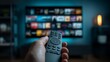 © Rifqi - Person's hand operating a remote control in front of a blurred TV screen showing numerous streaming options, concept for home entertainment, modern technology and digital media consumption