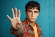 © KyleAnderson - Young man with curly hair and blue eyes wearing a red and orange plaid shirt over a yellow t-shirt extending his hand towards the camera with a serious expression against a blue background