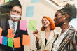 © artitwpd - Diverse coworkers brainstorming with colorful sticky notes on a glass wall in a modern office, smiling and collaborating during a creative planning meeting.