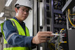© Sumala - industrial maintenance, workplace safety, Male engineer in safety helmet and reflective vest inspecting electrical control panel with tablet and walkie-talkie, modern energy infrastructure