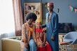 © Seventyfour - Black woman, Black man, and Black girl wearing traditional clothing celebrating Kwanzaa, girl sitting between adults, all facing camera with neutral expressions in living room