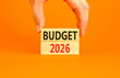 © Dzmitry - Budget 2026 symbol. Concept words Budget 2026 on beautiful wooden blocks. Beautiful orange paper background. Businessman's hand. Business and budget 2026 concept. Copy space.