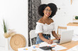 © etonastenka - Portrait of African American woman smiling and looking at camera while sitting at desk with laptop in bright modern apartment. Concept of remote work, online communication, and positive modern