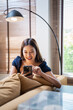 © Fahng - Happy young asian woman checking social media holding smartphone at home. Smiling young  woman using mobile phone app playing game, shopping online, ordering delivery relax on sofa.