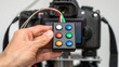 © ng-TDRD - A hand holding a remote control with colored buttons in front of a camera, suggesting functionality for photography or videography.