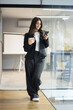 © Panorama - A smiling young businesswoman stands in a modern office, holding a coffee mug while looking at her smartphone