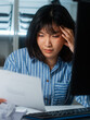© SAT65 - Serious Young Asian Businesswoman reading financial report document, reviewing data and information for making decision, planning business strategy feeling thought and stress in corporate office night