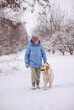 © StockMediaSeller - Elderly woman in a blue winter coat walking her Golden Retriever along a snowy rural path. She looks at the dog affectionately as they walk together surrounded by winter trees and fresh snow.