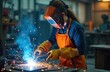 © Viktor - Young woman welds metal parts in busy industrial workshop. Wears protective mask, gloves, orange apron for safety. Bright sparks, blue smoke fly from metal joint. Female worker operates machine,