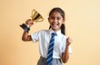 © Viktor - Young Indian schoolgirl in uniform holding golden trophy and smiling. Happy female student with victory cup on beige background. Cheerful teenage girl celebrating success in education.