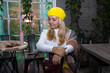 © Светлана Высокос - A woman with long blonde hair, dressed in a yellow beret and a white sweater, is sitting at a table in a cozy, autumnal cafe.