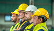 © BESTIMAGE - A group of construction workers wearing hard hats and safety vests are standing in a row looking at something.