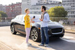 © sofiko14 - A woman and a man shaking hands next to a black electric car, possibly after a deal or agreement. The man is holding a phone, and the woman is holding a coffee cup.