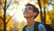 © miss irine - Smiling teenage boy with glasses wears backpack, looks up with hope in sunny park. He is ready for education, travel, and self-discovery in autumn.