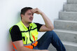 © eakgrungenerd - Construction worker in safety vest sitting on stairway with hands on head, showing signs of stress, fatigue, or headache. Concept of burnout, job pressure, mental health, and labor exhaustion.