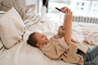 © BullRun - Happy girl lying in bed smiling at phone, joyful interaction with digital content showing relaxed modern childhood and comfort in cozy home environment.