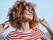 © SHOTPRIME STUDIO - Young woman with curly hair smiles in sunglasses and a striped shirt outdoors, capturing carefree summer style and radiant confidence against a bright blue sky.