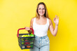 © luismolinero - Young caucasian woman holding a shopping basket full of food isolated on yellow background showing ok sign with fingers