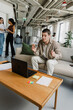 © Drobot Dean - A male worker sits on a couch and looks at a laptop on a table while two female workers walk and talk next to him