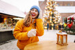 © maxbelchenko - Smiling woman in yellow jacket holds hot drink at bustling winter market. Beautiful tourist enjoys New Year's atmosphere at fair decorated with lights, holding mulled wine. Concept is festive, cozy.