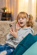 © Andrii Iemelianenko - Young teenager girl sits at home scrolling pictures on a smartphone, laughing at funny memories. Blonde child kid on sofa points at the screen, chatting about family adventures with big smiles.