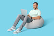 © Pixel-Shot - Stylish young man in jeans with modern laptop sitting on beanbag chair against blue background