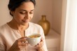 © SERHII - A mature South Asian woman relaxing while holding a cup of herbal chamomile tea. A quiet moment of self-care and wellness at home. Close-up with soft natural light