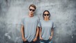 © Tetiana - Stylish Hipster Couple In Blank Gray T-Shirts, Jeans, And Sunglasses Poses Against Concrete Wall In Urban City Street - Front Tshirt Mockup.