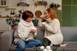 © Sunny - Two women enjoying warm beverages while sitting on a cozy sofa, wearing knitted sweaters, surrounded by festive decorations and a warm, inviting atmosphere for a joyful gathering