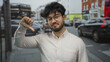 © Krakenimages.com - Young man with beard showing thumbs down gesture outdoors on busy street with cars and buses in background, wearing glasses and white shirt in urban setting.