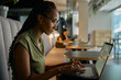 © StratfordProductions - Side view of focused young black businesswoman typing on laptop sitting at desk in office lounge
