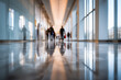 © Daria - Blurred silhouettes of people walking through a brightly lit modern corridor with large windows reflecting on polished marble floors during daytime