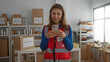 © Krakenimages.com - Young woman volunteer using phone in charity donations center room with boxes in the background showcasing a community support environment indoors.
